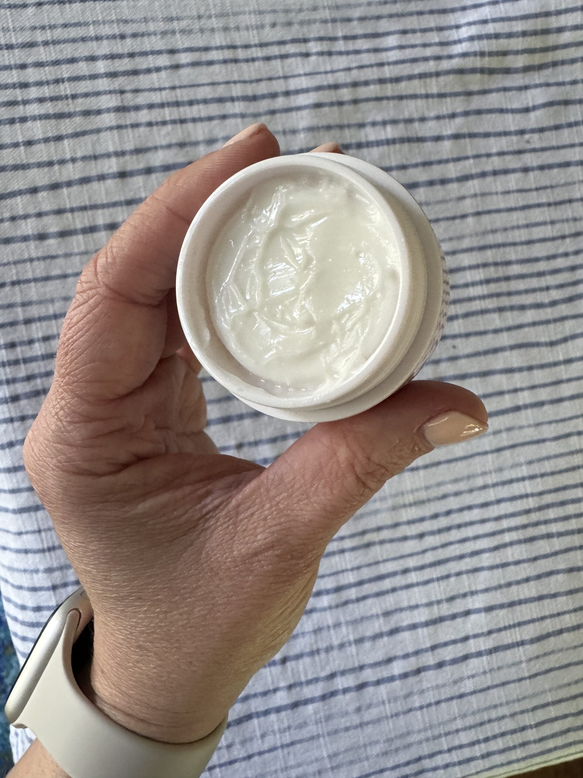 A hand holding an open jar of white cream against a striped fabric background.