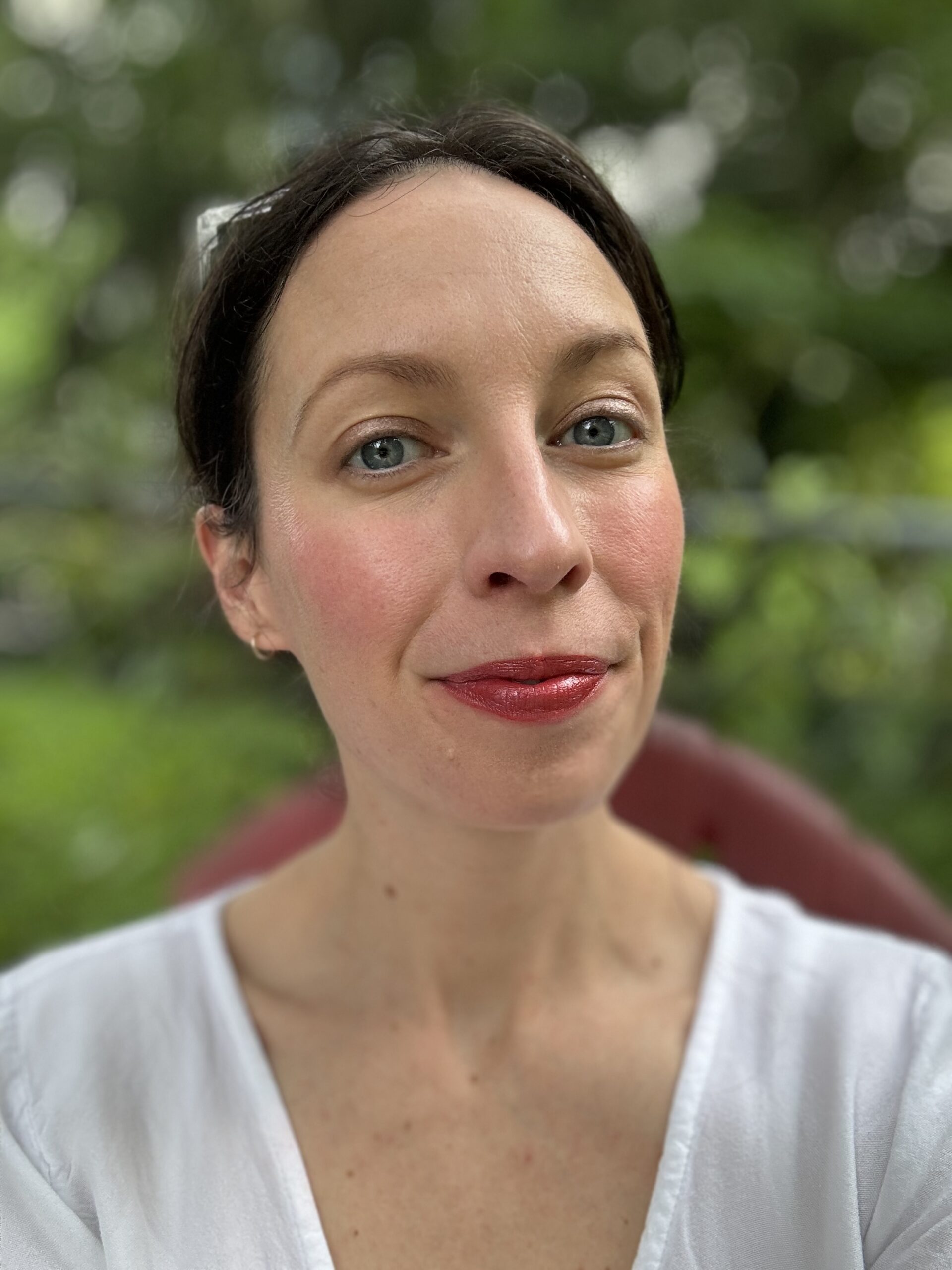 A woman with light skin, dark hair, and blue eyes is wearing red lipstick and a white shirt, posing against an outdoor background.