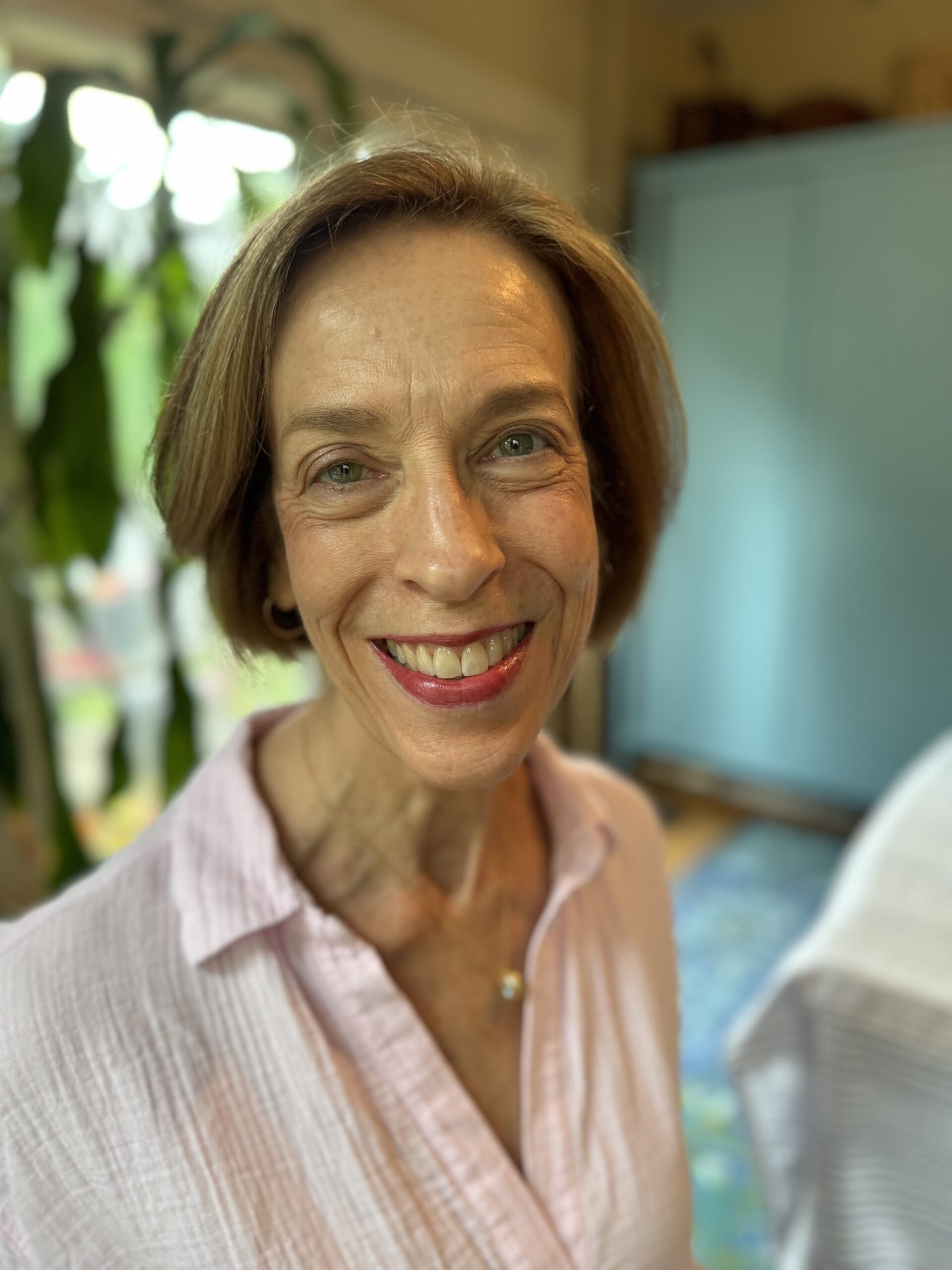 A person with short brown hair and a light pink shirt smiles at the camera in a room with a plant and blue cabinet in the background.