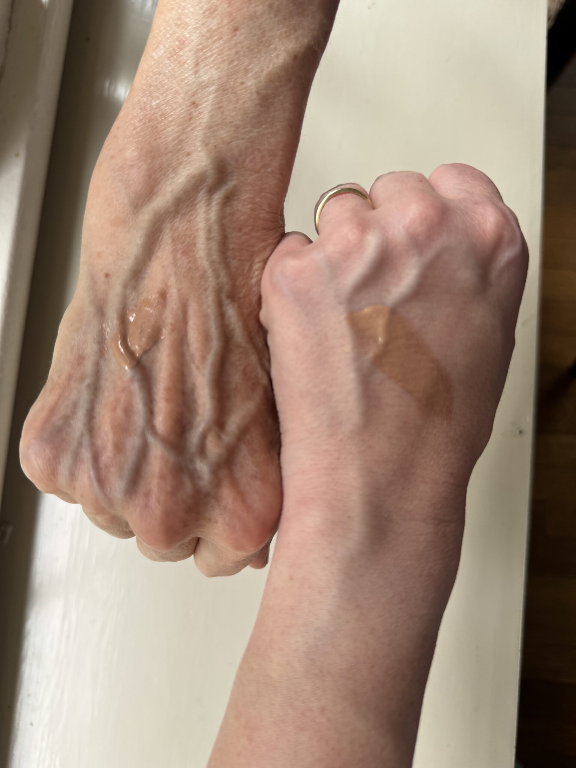 Two hands, one older and one younger, are seen with patches of skincare product applied on the backs. The hands are positioned side by side on a light-colored surface.
