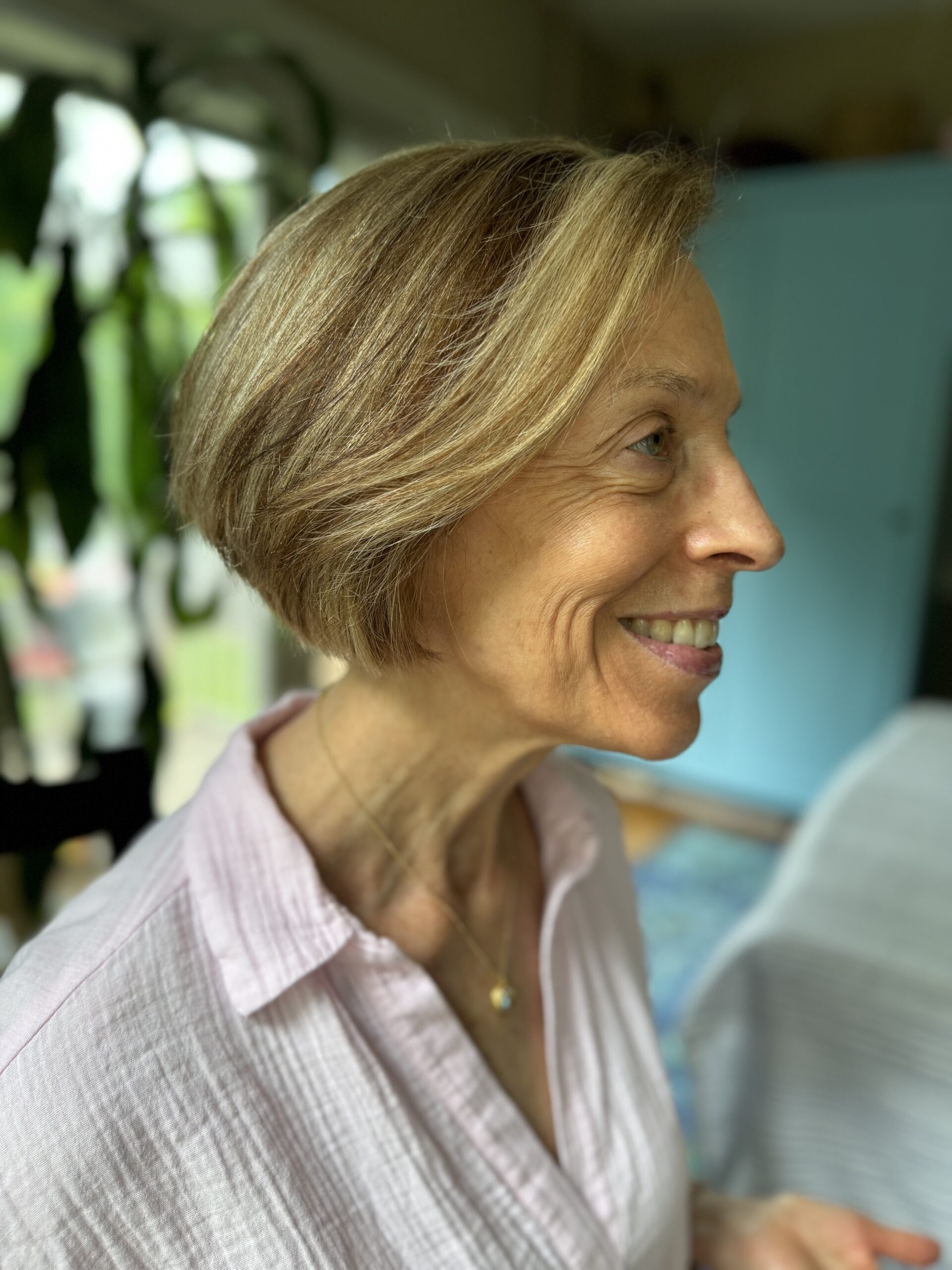 A woman with short blonde hair and a light pink shirt smiles while looking to the side. There is a blue piece of furniture and green plants in the background.