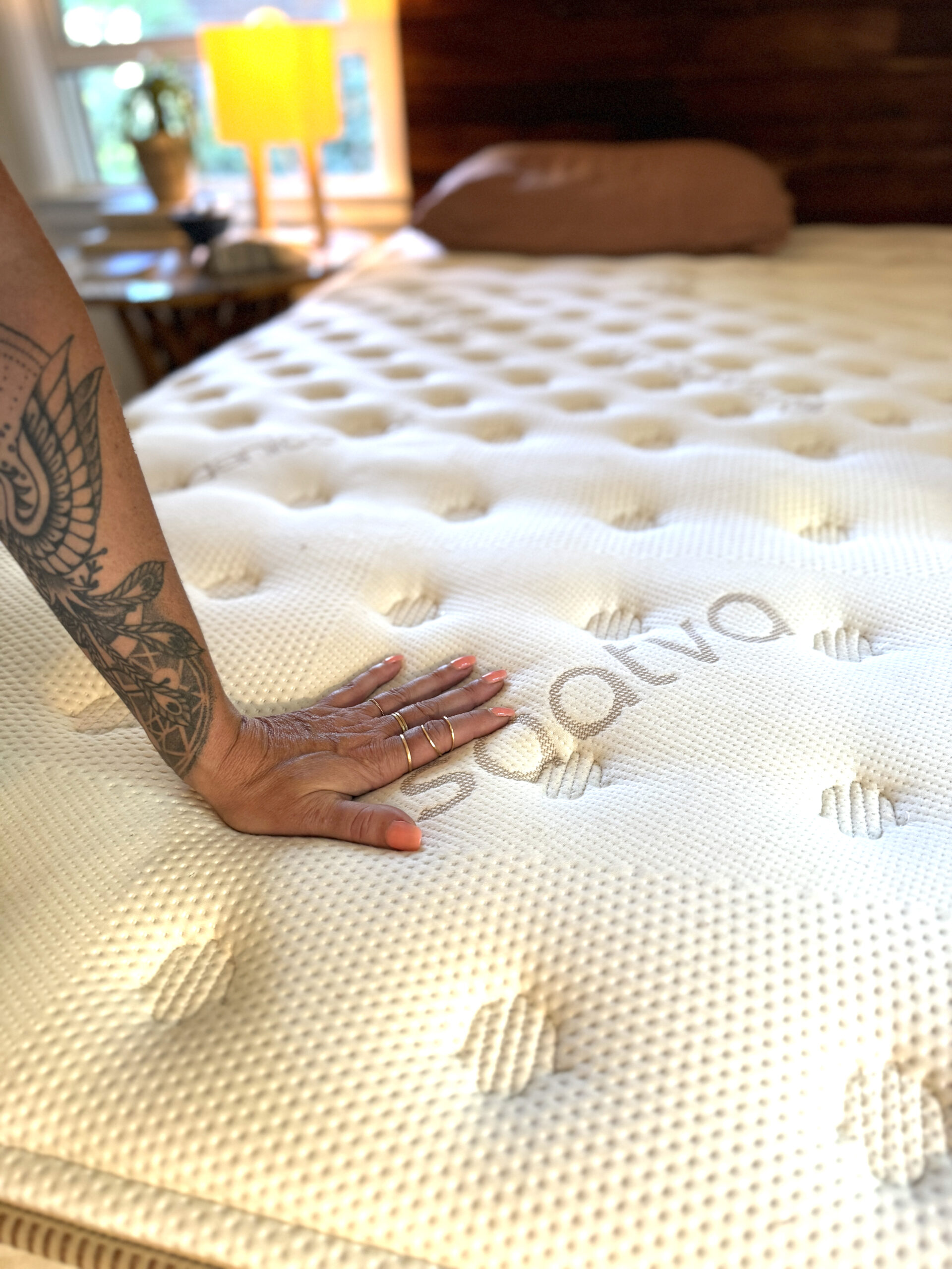 Reviewer Trinity Wilbourn's hand presses down on a Saatva nontoxic mattress in a sunlit bedroom, showcasing its comfort and quality.