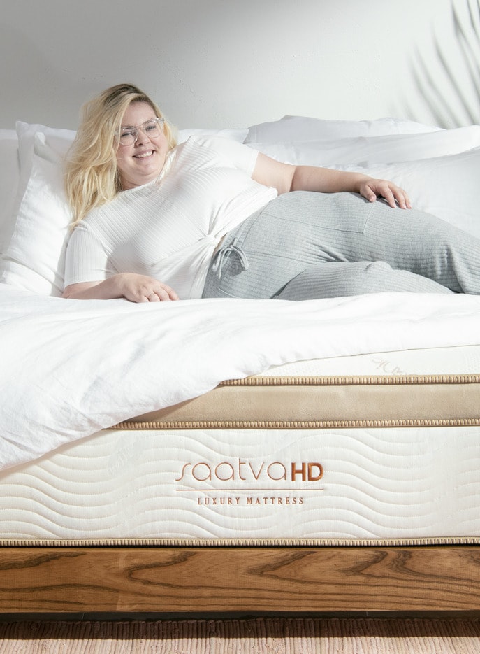 A person reclines on a bed with white sheets, enjoying the comfort of a Saatva HD luxury organic mattress.