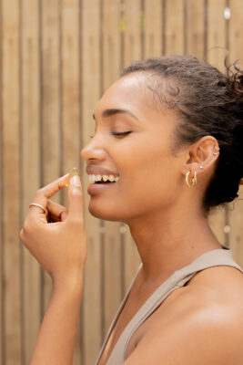 A woman in a sleeveless top holds a yellow capsule near her mouth, smiling, with a wooden fence background.