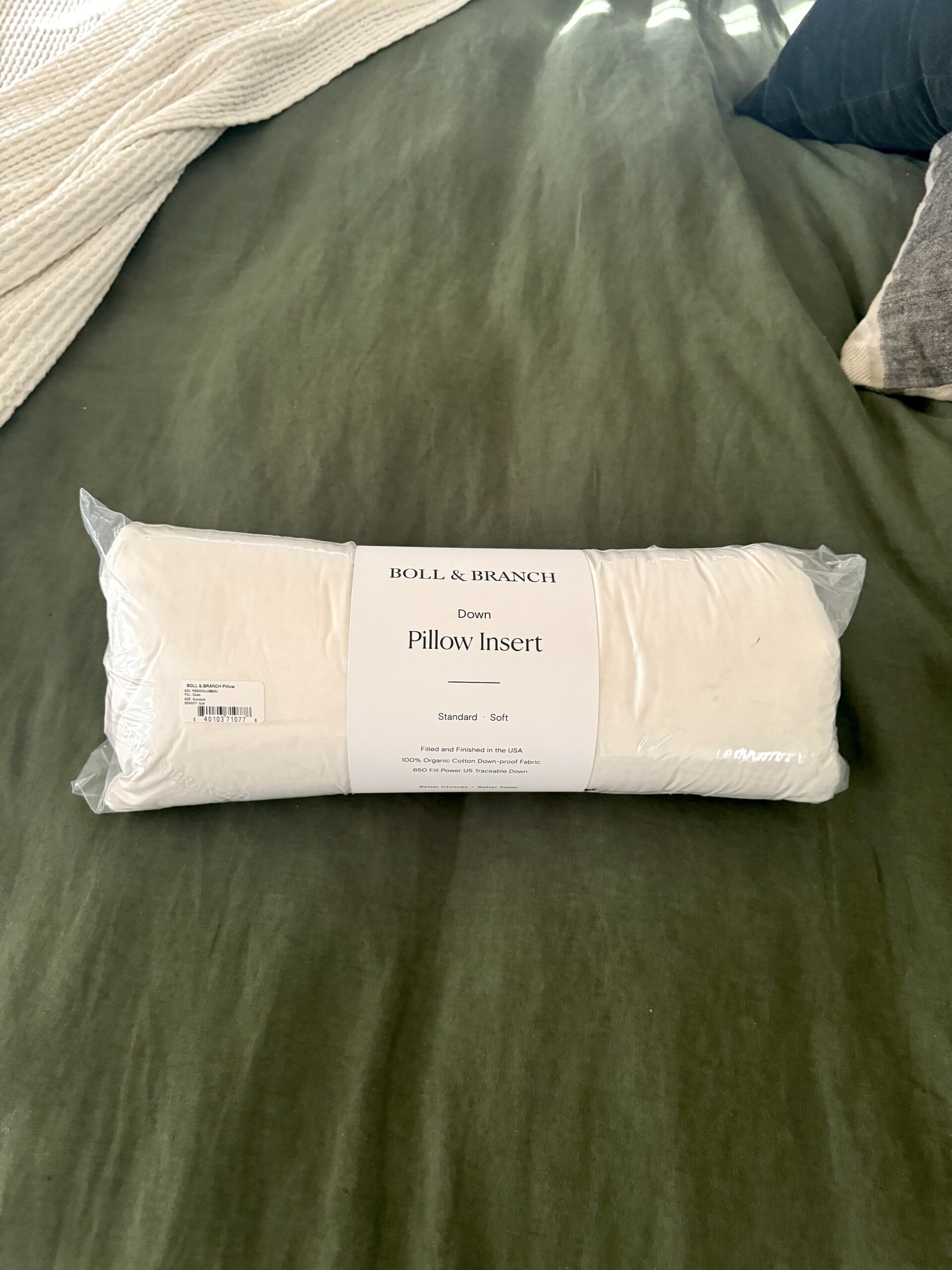 A down pillow insert, packaged in plastic with a label reading "Boll & Branch Down Pillow Insert Standard Soft," is placed on a green bedspread.