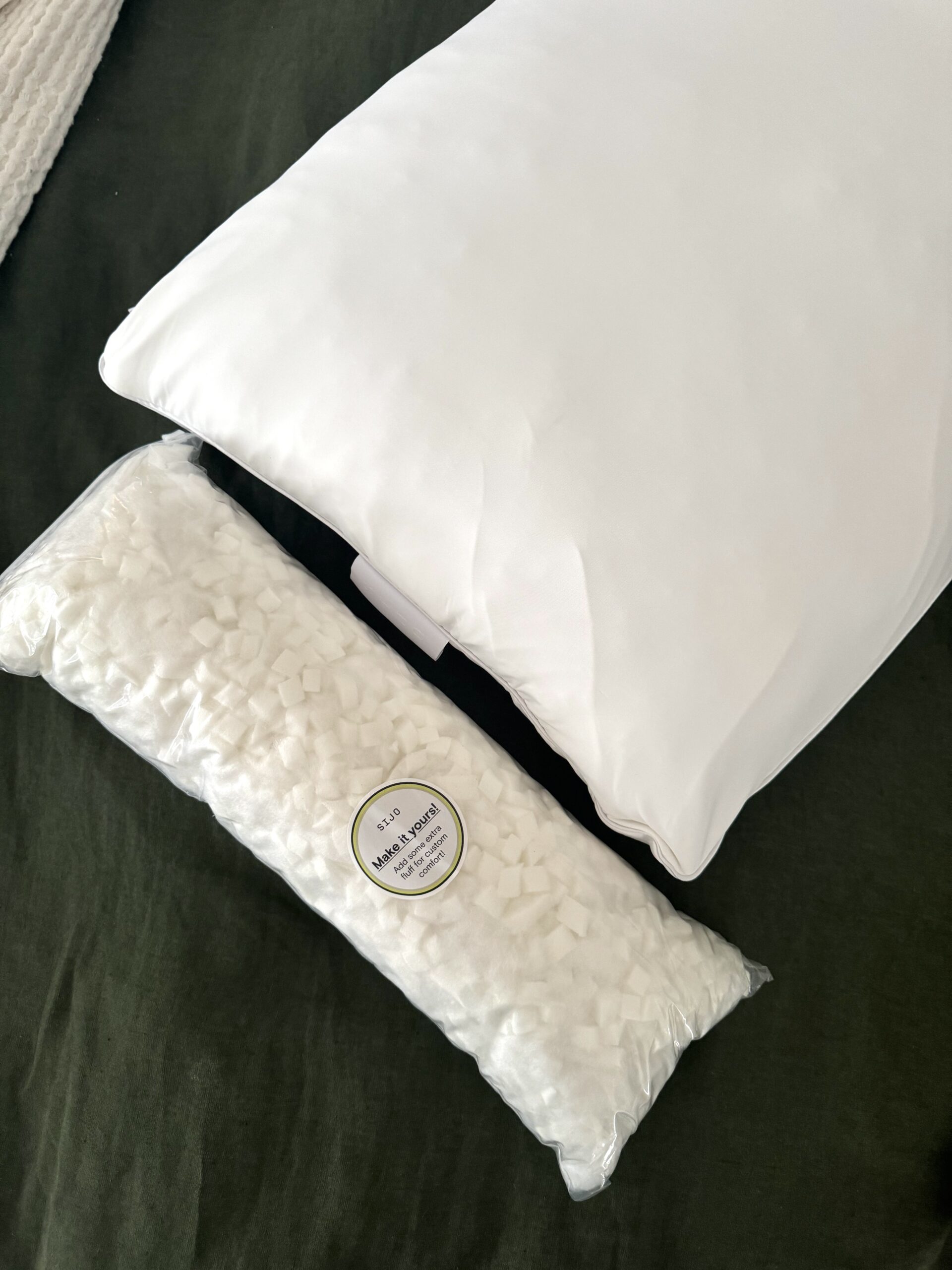 A white pillow is placed on a dark green surface next to a clear plastic bag filled with small, white foam cubes.