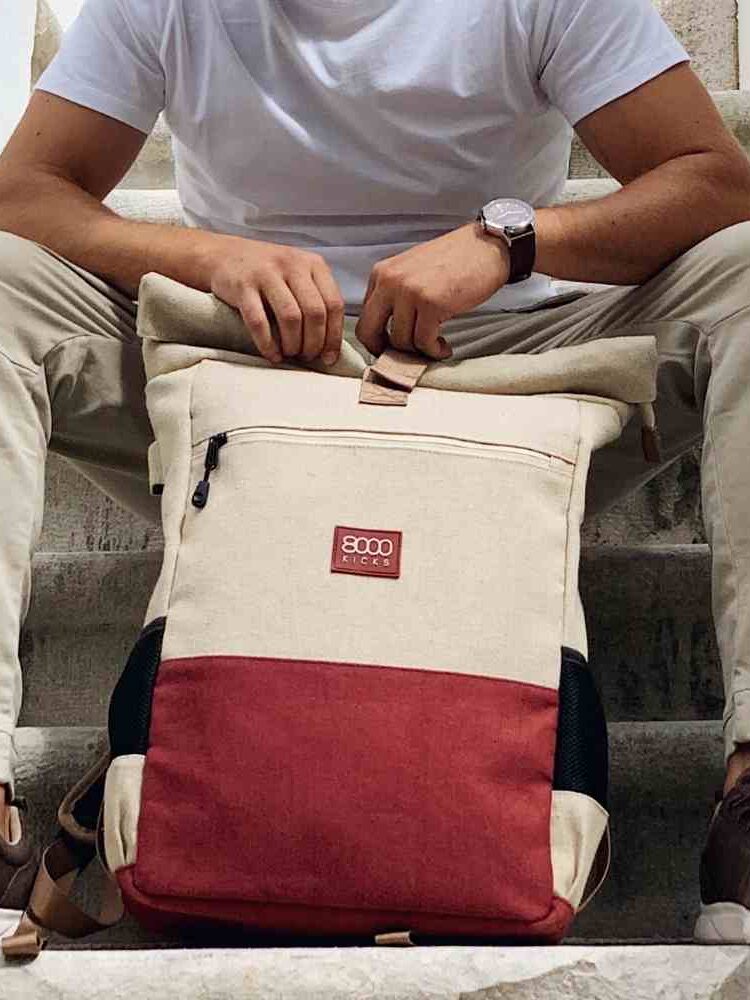 Person in neutral clothing sits on stairs holding a beige and red backpack with a visible front zipper and logo patch.
