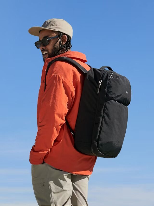 A man wearing sunglasses, a tan cap, an orange jacket, and khaki pants stands outdoors with a black backpack against a clear blue sky.