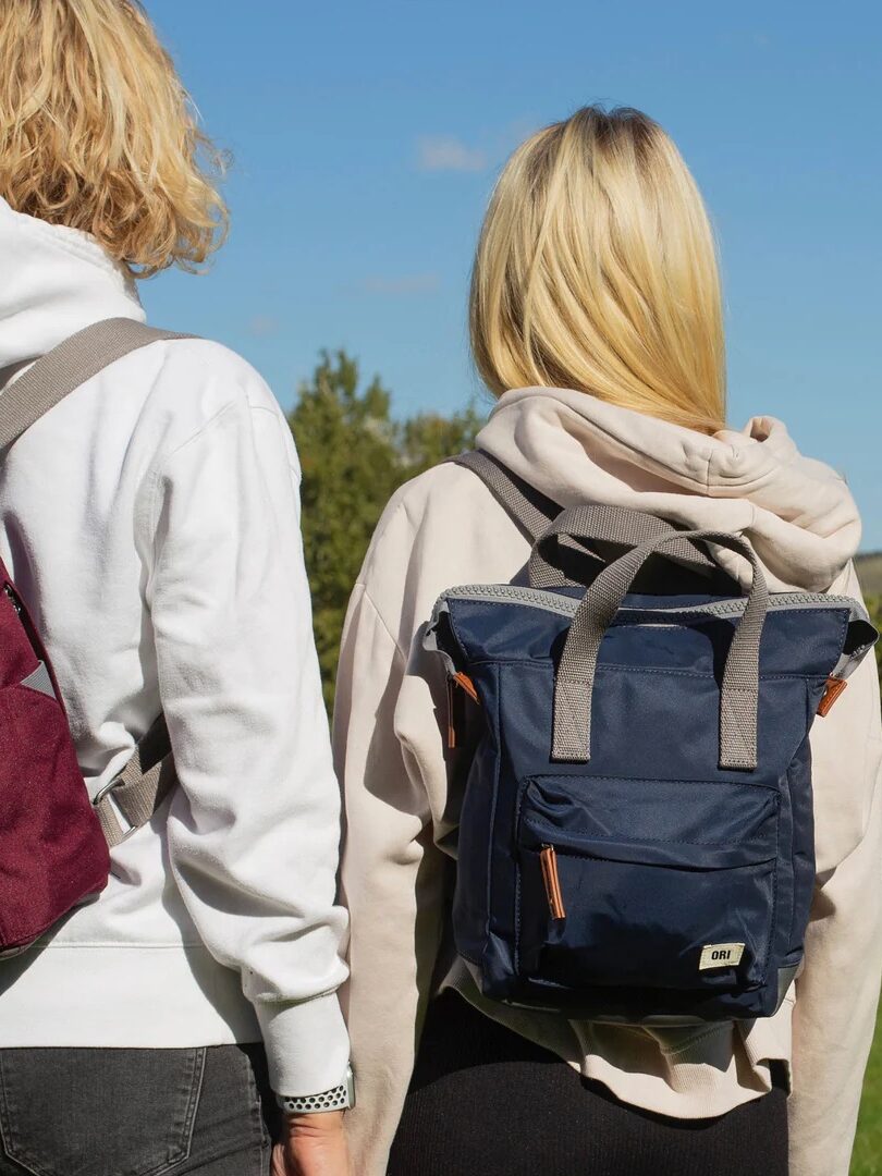 Two people wearing backpacks and hoodies stand outdoors facing away, with greenery and blue sky in the background.