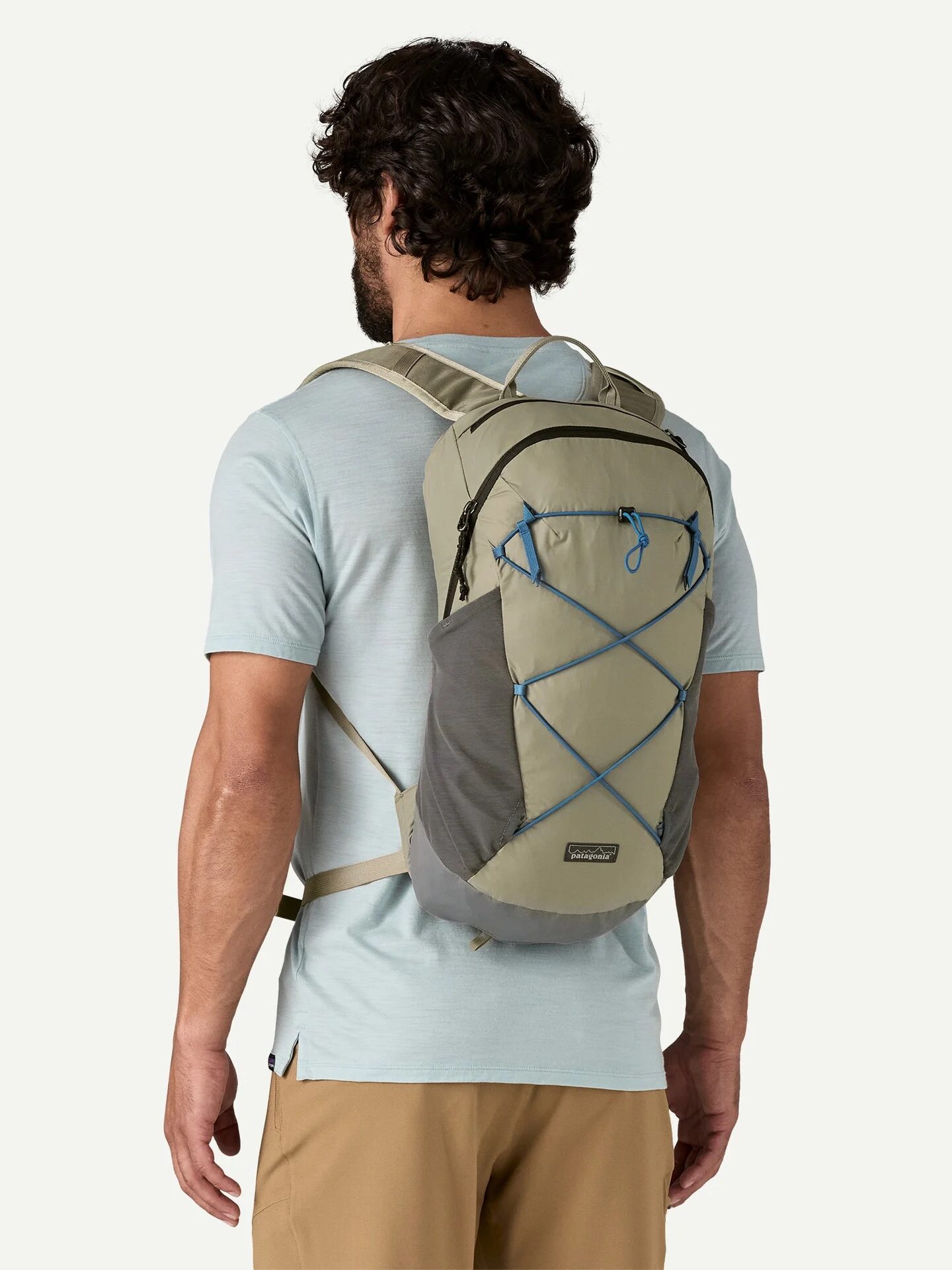 A man wearing a light blue t-shirt and tan pants stands facing away, modeling a gray and beige hiking backpack with blue drawstrings.