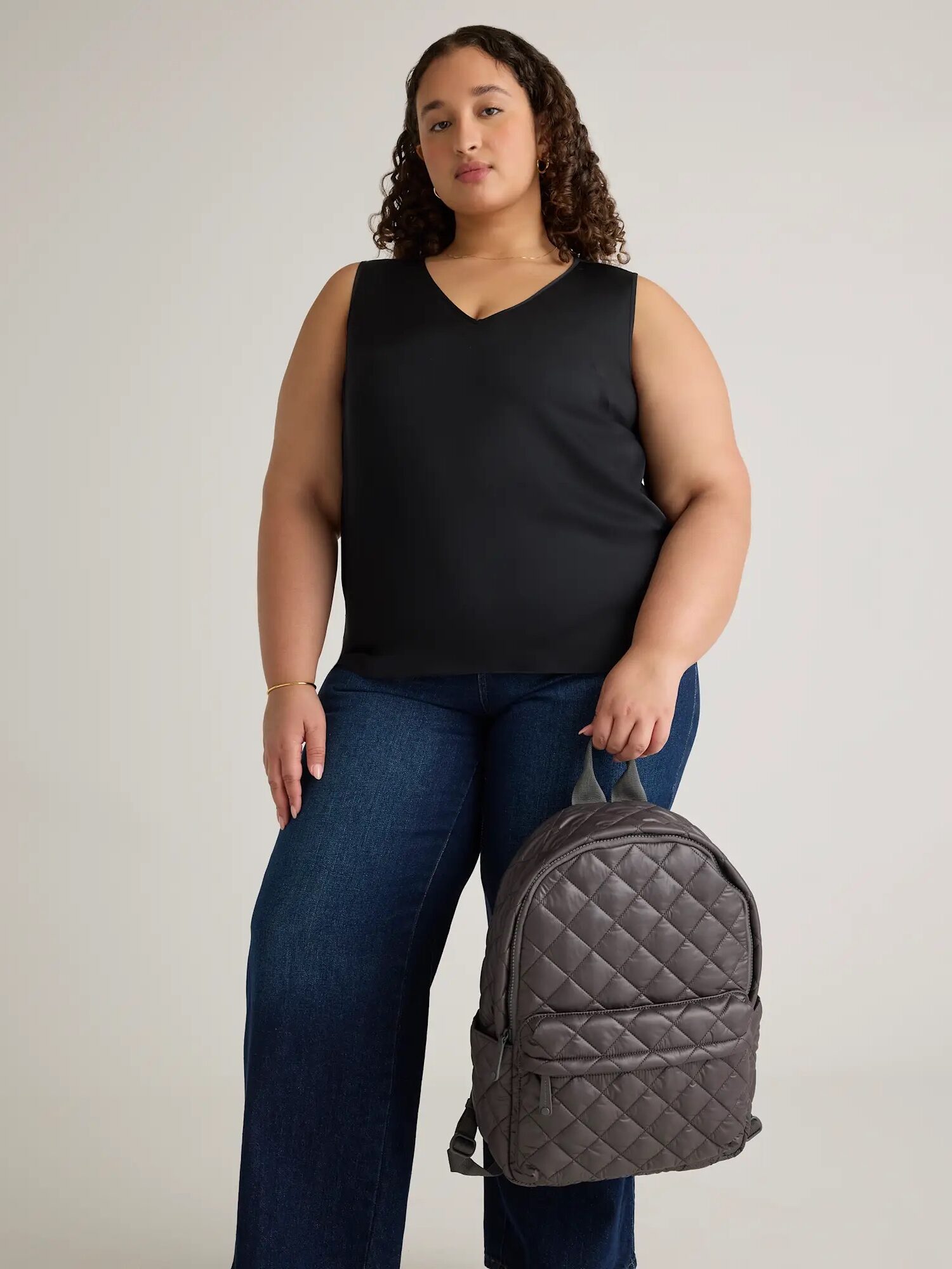 A woman wearing a black sleeveless top and blue jeans stands holding a quilted gray backpack in her left hand against a plain background.