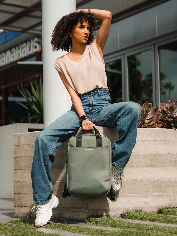 A woman in a sleeveless beige top and blue jeans sits on a low concrete wall, holding a gray backpack, with a modern building and plants in the background.