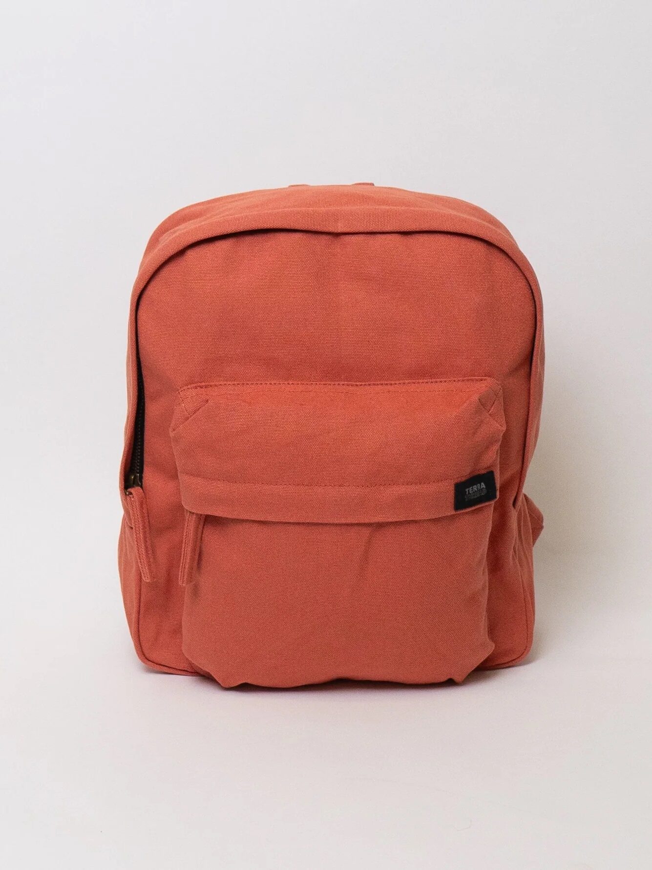 A plain, coral-colored backpack with a front zip pocket and a small black label, photographed against a white background.