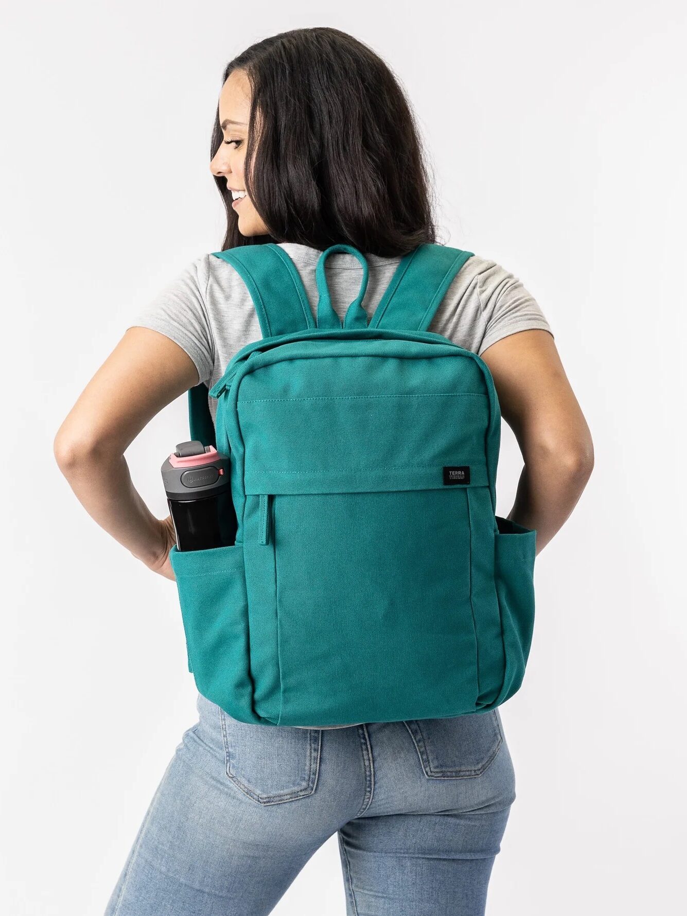 Person wearing a teal backpack with side pockets, holding a water bottle in one pocket, facing away and smiling over their shoulder against a plain white background.