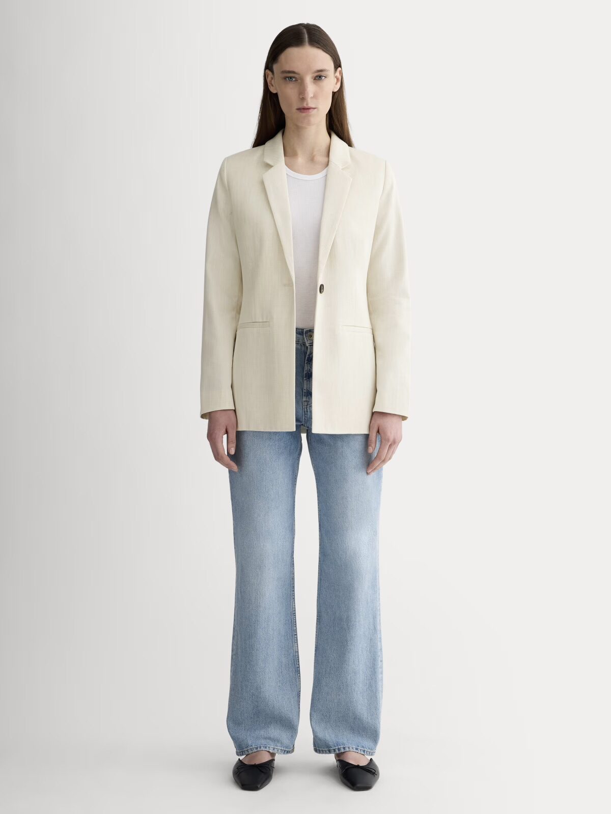 A woman stands facing forward, wearing a cream blazer over a white top, light blue wide-leg jeans, and black shoes, against a plain light background.