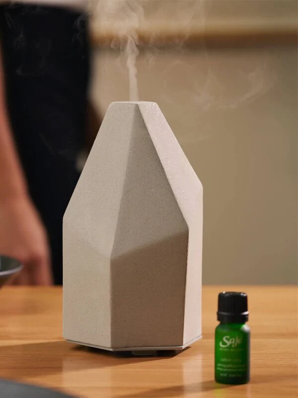 Geometric diffuser emitting vapor on a wooden table with essential oil bottle and bowl of figs nearby. Person in the background.