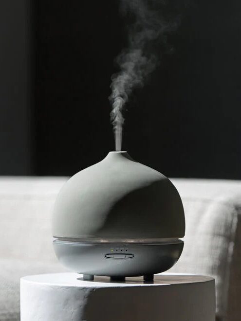 A small, gray, dome-shaped aroma diffuser emits steam while placed on a white surface, with a neutral background.