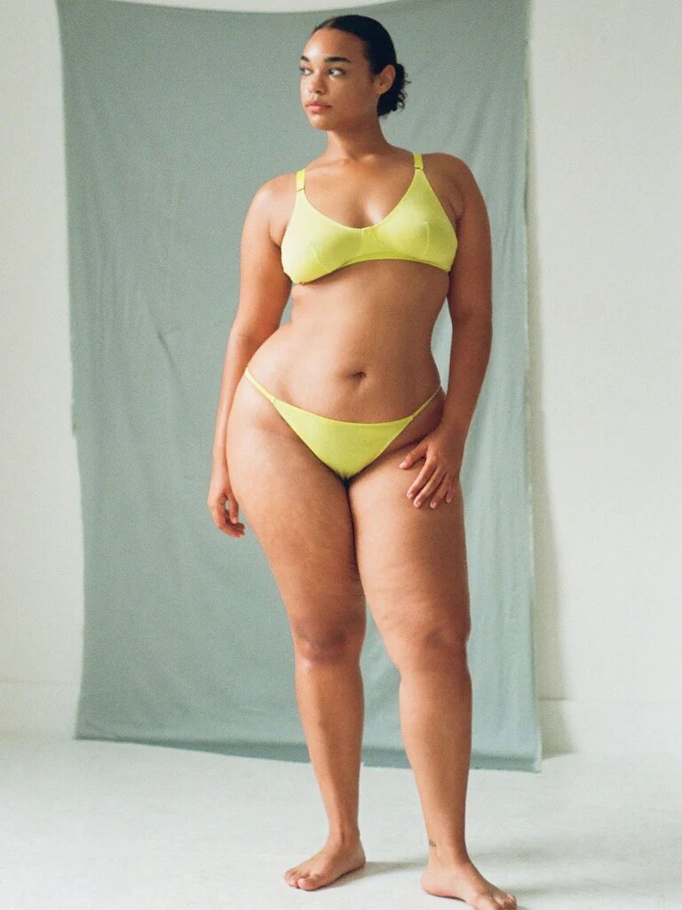 Person wearing a lime green bikini stands against a light gray backdrop in a studio setting.