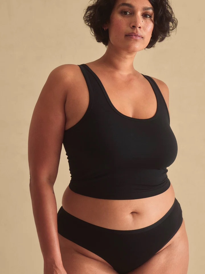 A person wearing a black tank top and black underwear stands against a plain beige background.