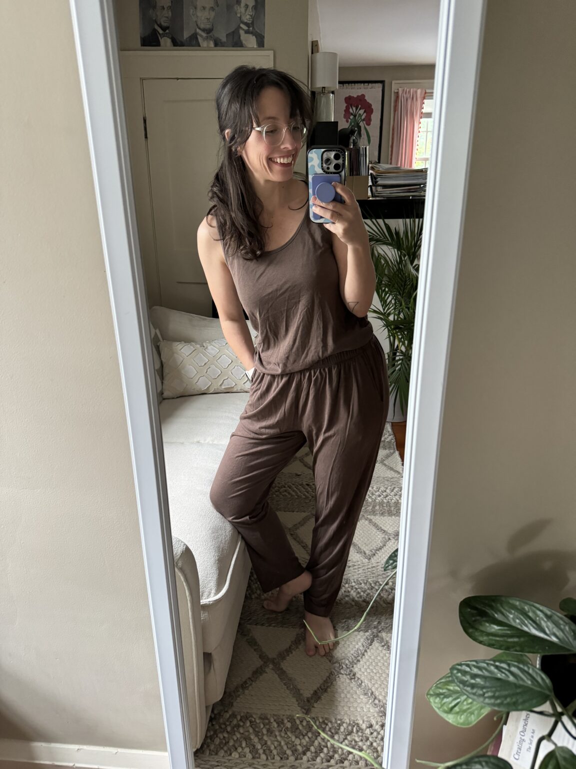 A woman wearing brown loungewear stands barefoot in front of a mirror, smiling and taking a selfie in a living room with plants and framed pictures in the background.