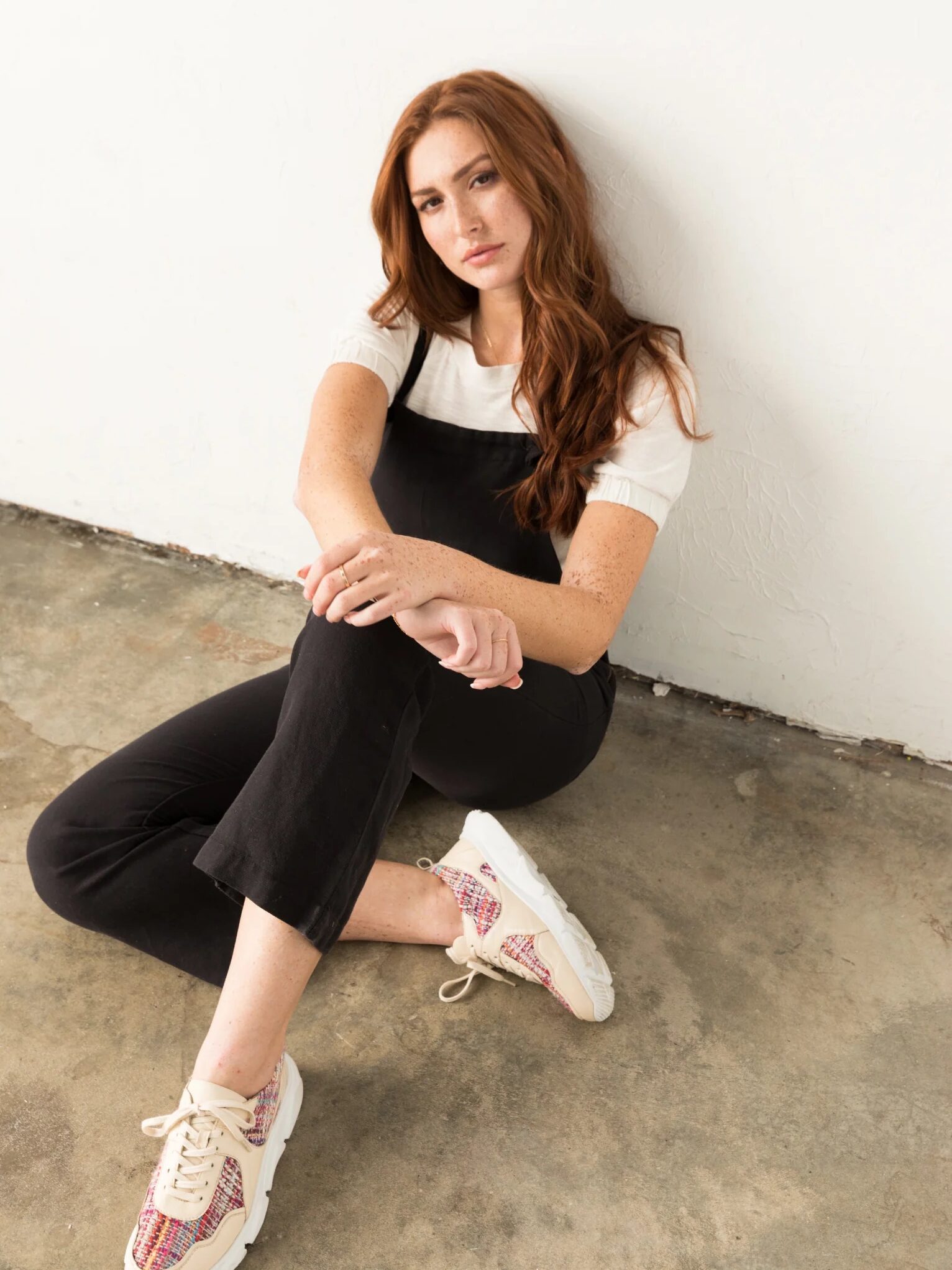 Person with long hair sits on the floor, leaning against a white wall. They are wearing a black outfit and colorful sneakers, looking towards the camera.