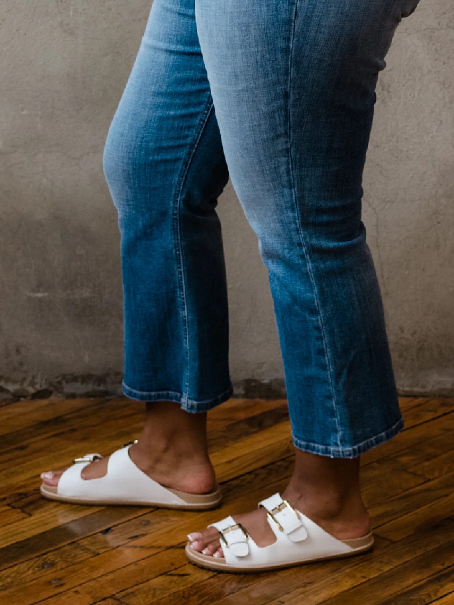 Person wearing blue jeans and white sandals stands on a wooden floor.