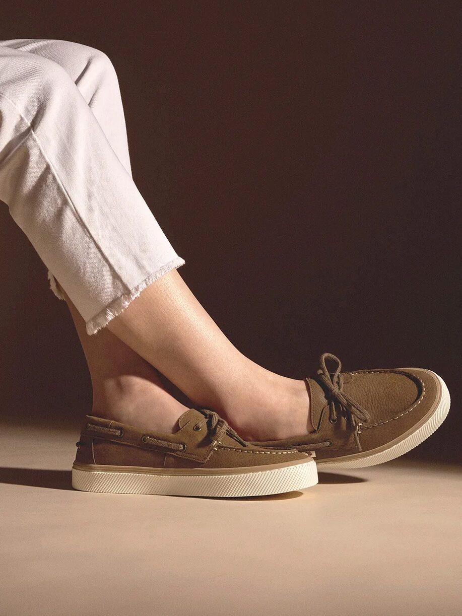 A person wearing brown boat shoes with white soles, paired with white pants, and a close-up of the same shoe on a wooden surface.