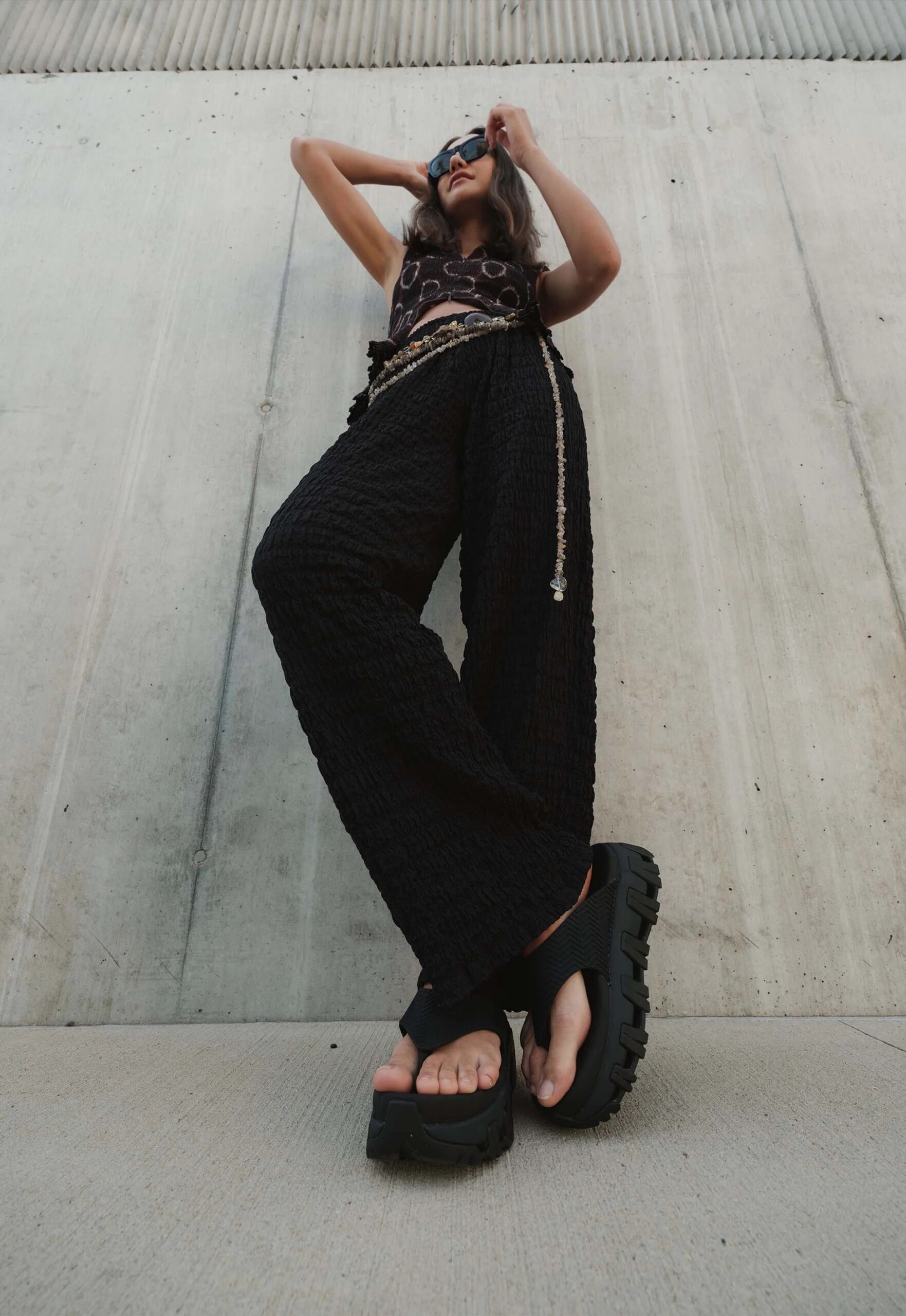 Person in black wide-leg pants and platform sandals stands against a concrete wall, adjusting sunglasses, viewed from a low angle.