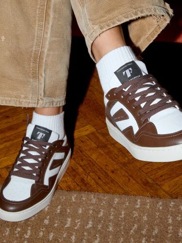 Person wearing brown and white sneakers with white socks and beige pants, standing on a wooden floor.