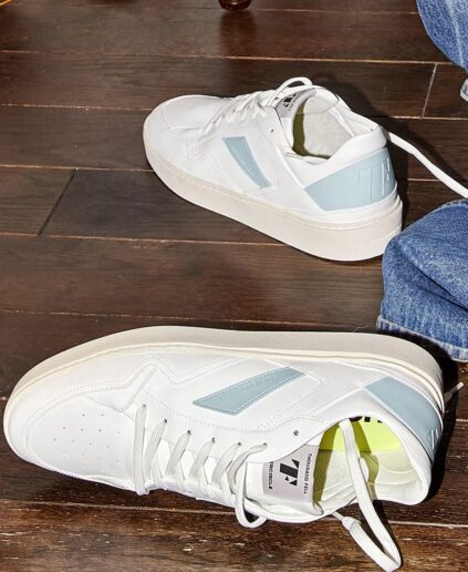 A pair of sustainable white low-top sneakers with light blue accents rest on a dark wooden floor next to a person wearing blue jeans.