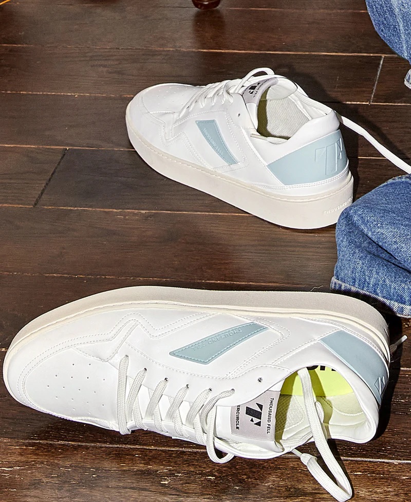 A pair of sustainable white low-top sneakers with light blue accents rest on a dark wooden floor next to a person wearing blue jeans.