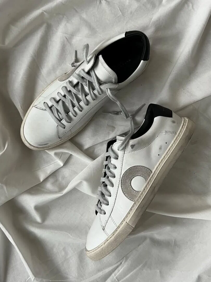 A pair of white sustainable sneakers with grey laces and black accents placed on a wrinkled white fabric background.