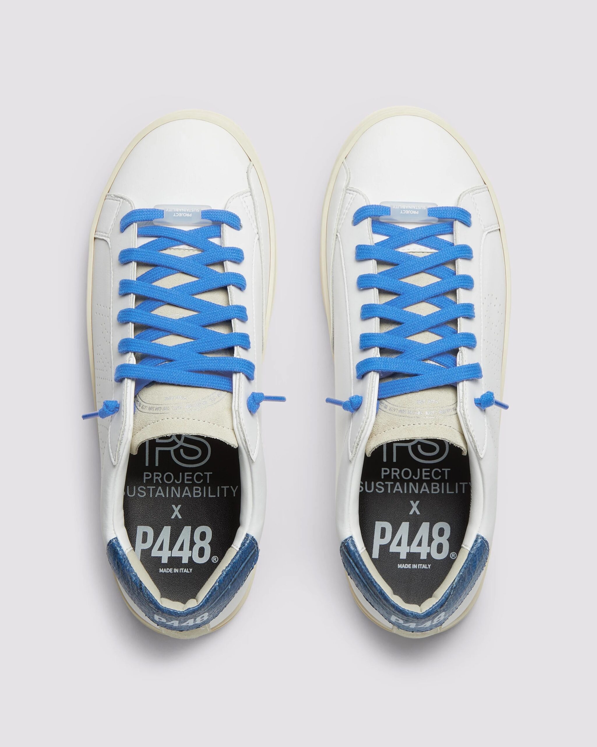 A pair of sustainable sneakers with white uppers, blue laces, black insoles labeled "PROJECT SUSTAINABILITY x P448," and blue heel accents are shown from a top-down view.