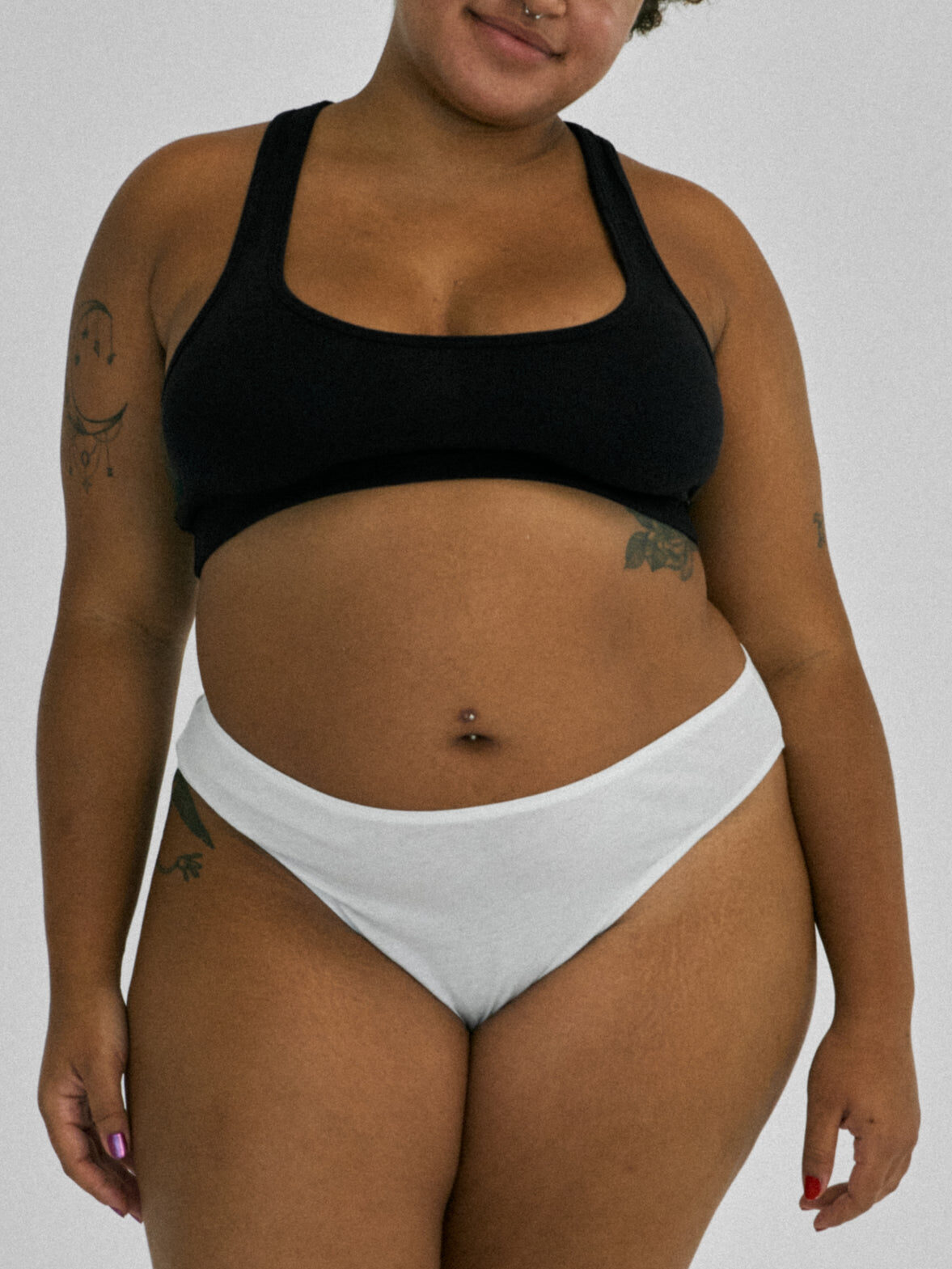 Person wearing a black sports bra and white underwear stands against a plain background, showing tattoos on their arms.