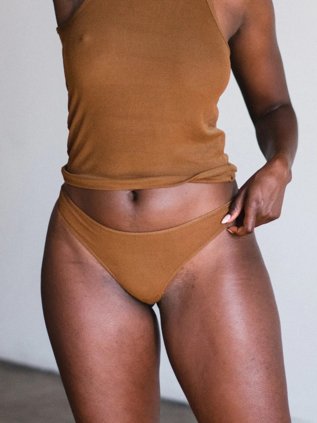 Person wearing a brown tank top and underwear, standing indoors against a plain background.