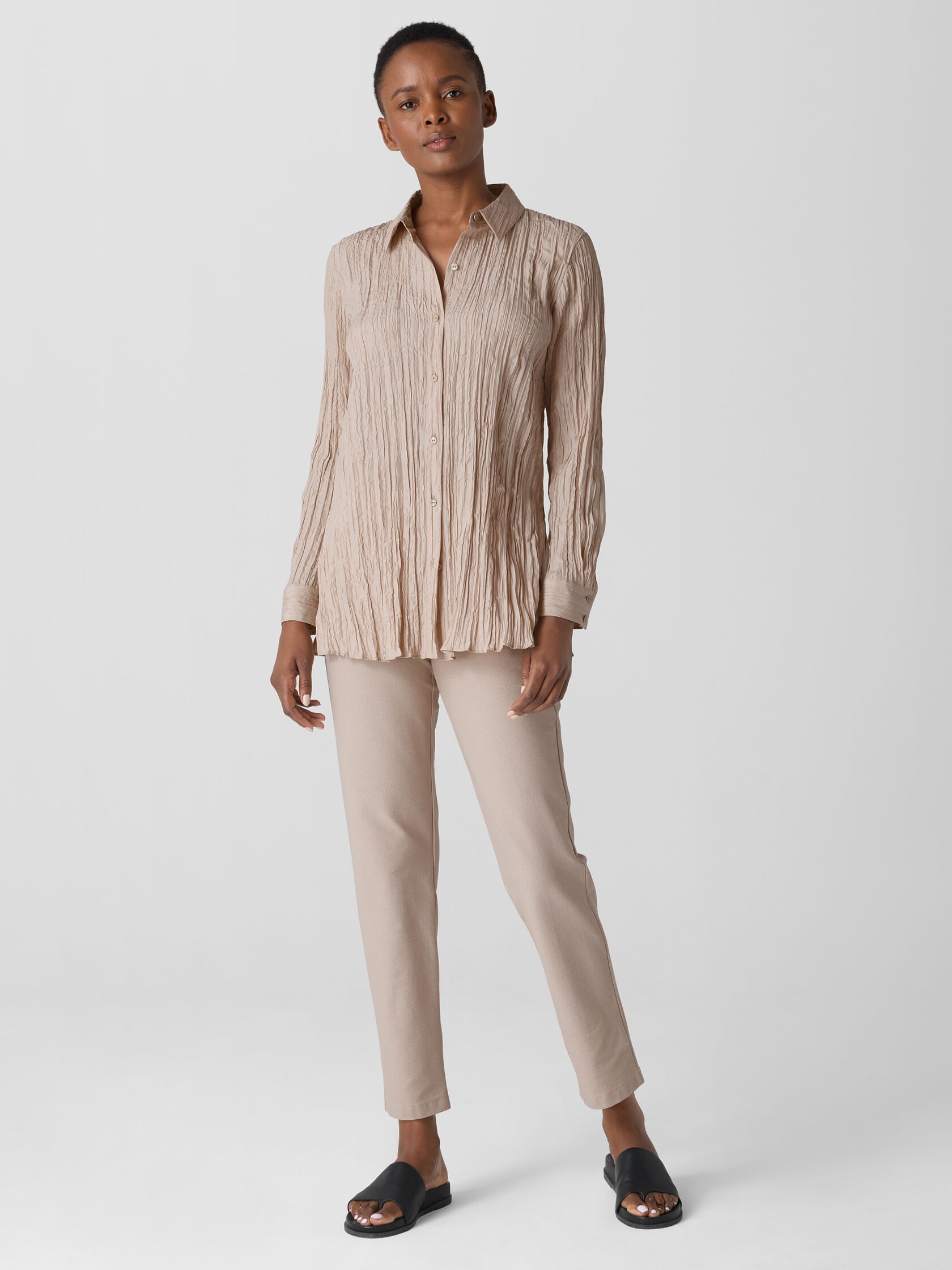 A person stands wearing a beige crinkled long-sleeve shirt, matching beige pants, and black slide sandals against a plain light background.