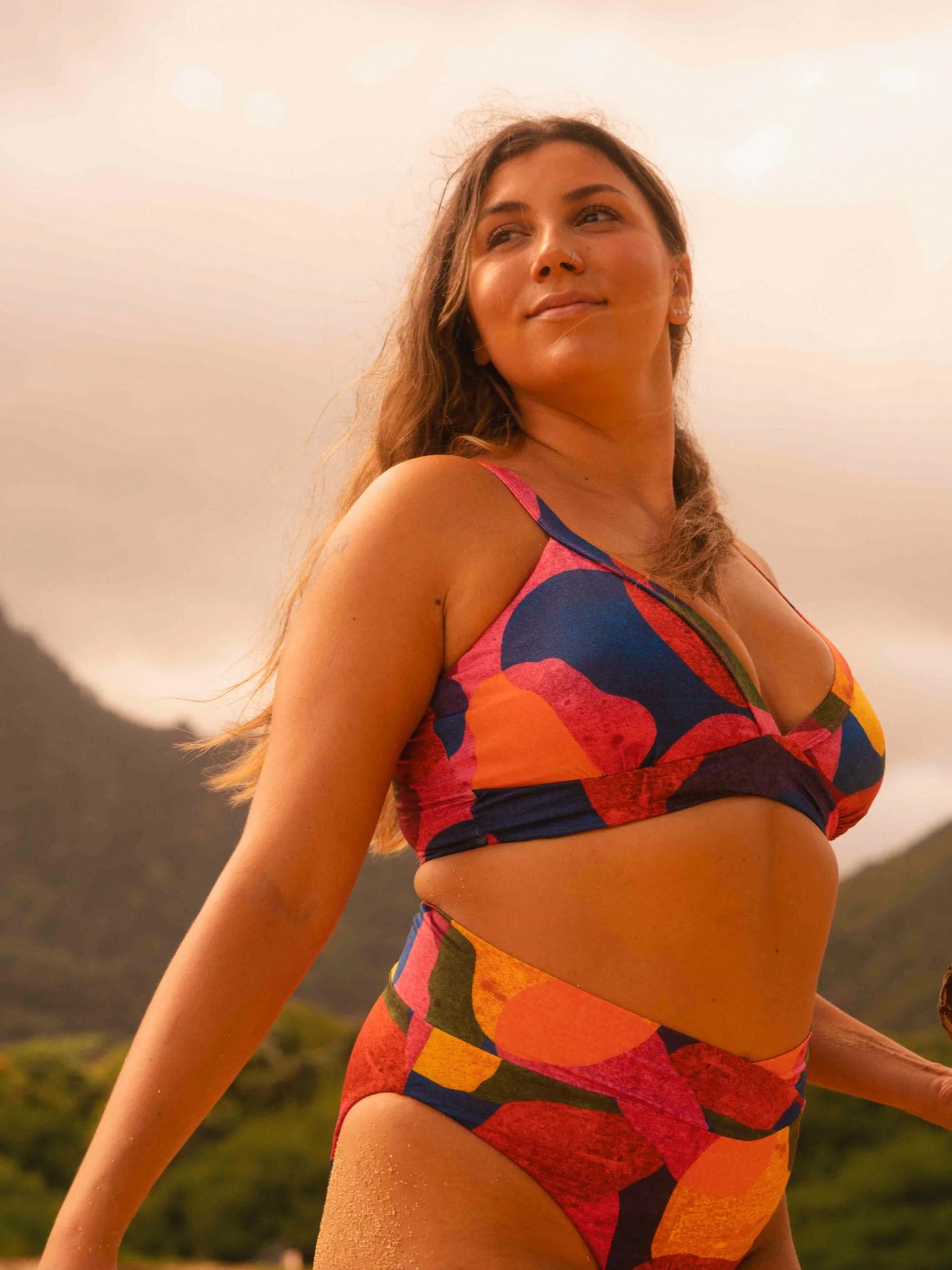 Person in colorful swimwear stands outdoors with mountains and cloudy sky in the background.