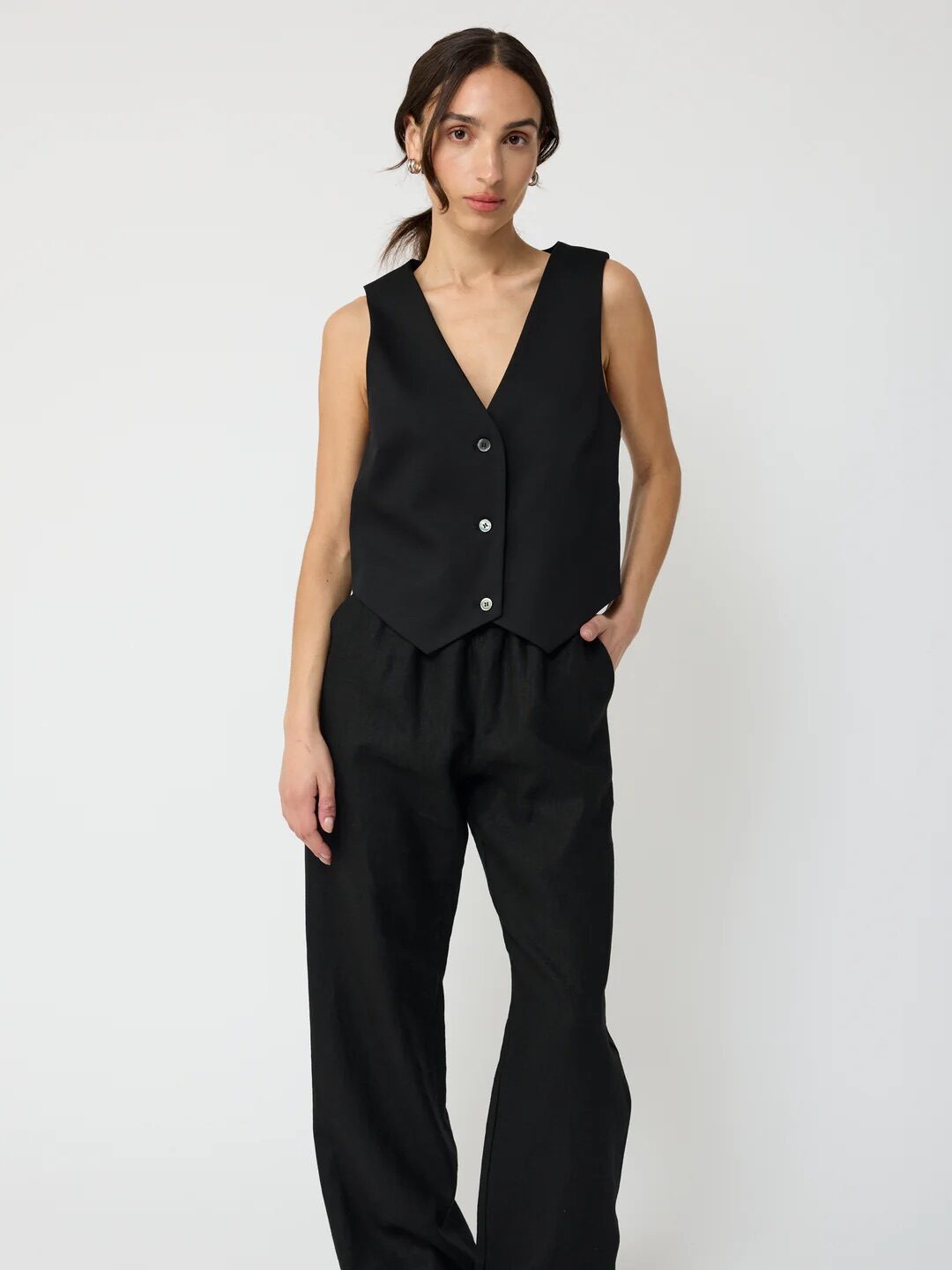 A woman stands against a plain background wearing a sleeveless black vest with buttons, loose black trousers, and black sandals.