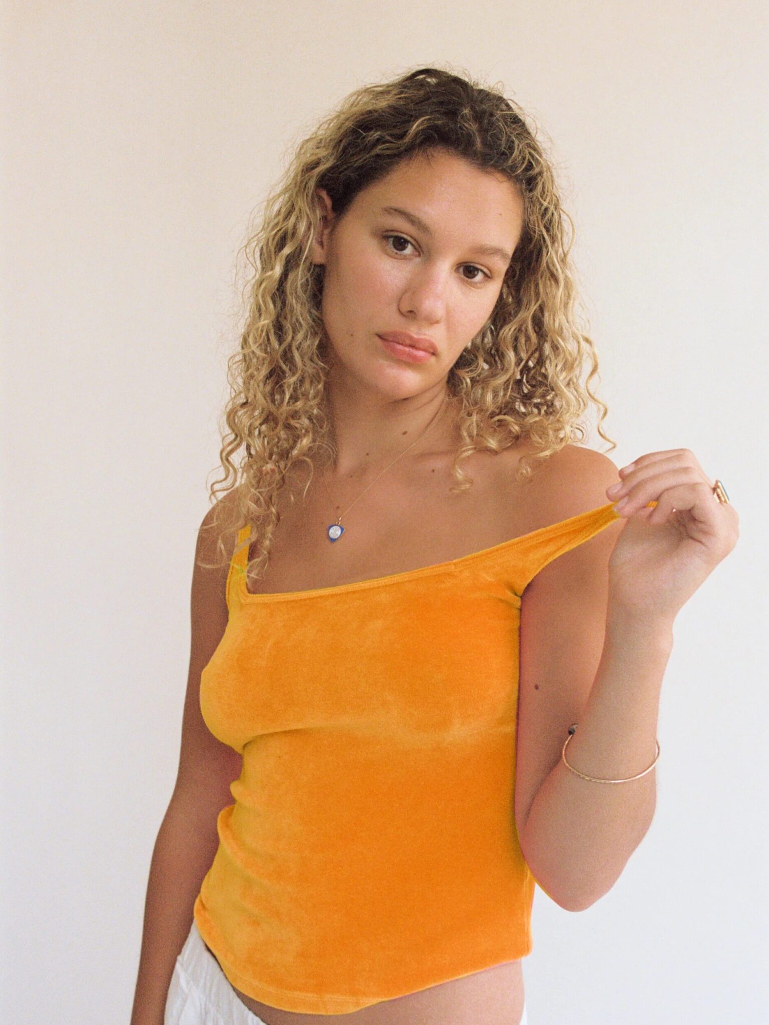 A woman with curly blonde hair wearing an orange off-shoulder top and white pants stands against a plain light background, looking at the camera.