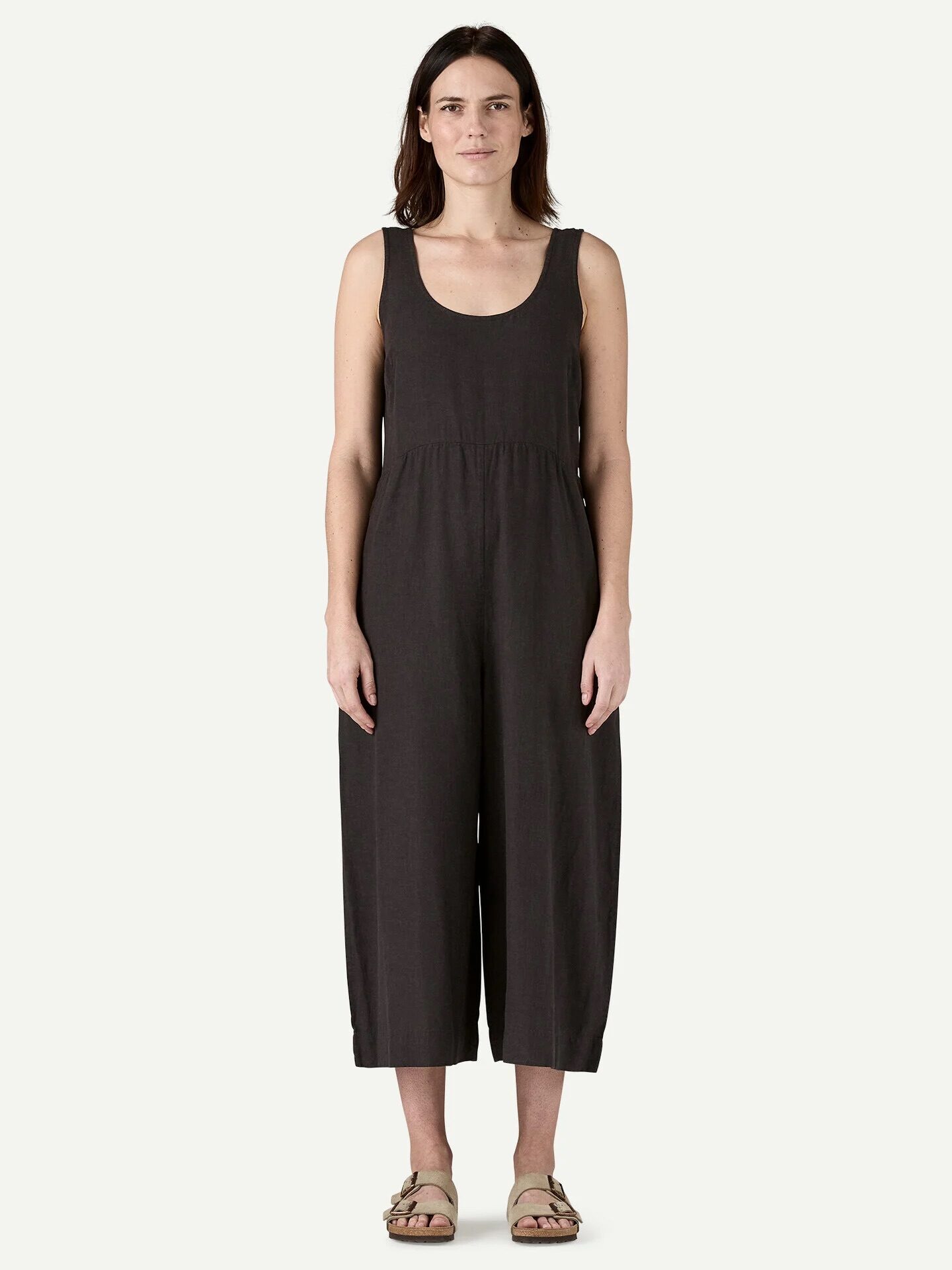 A woman stands facing forward, wearing a sleeveless black jumpsuit and beige sandals, against a plain light background.
