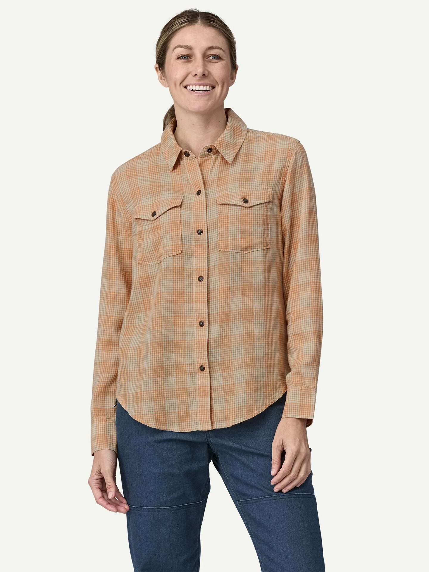 A woman wearing a peach and beige plaid button-up shirt and blue pants stands smiling against a plain light background.