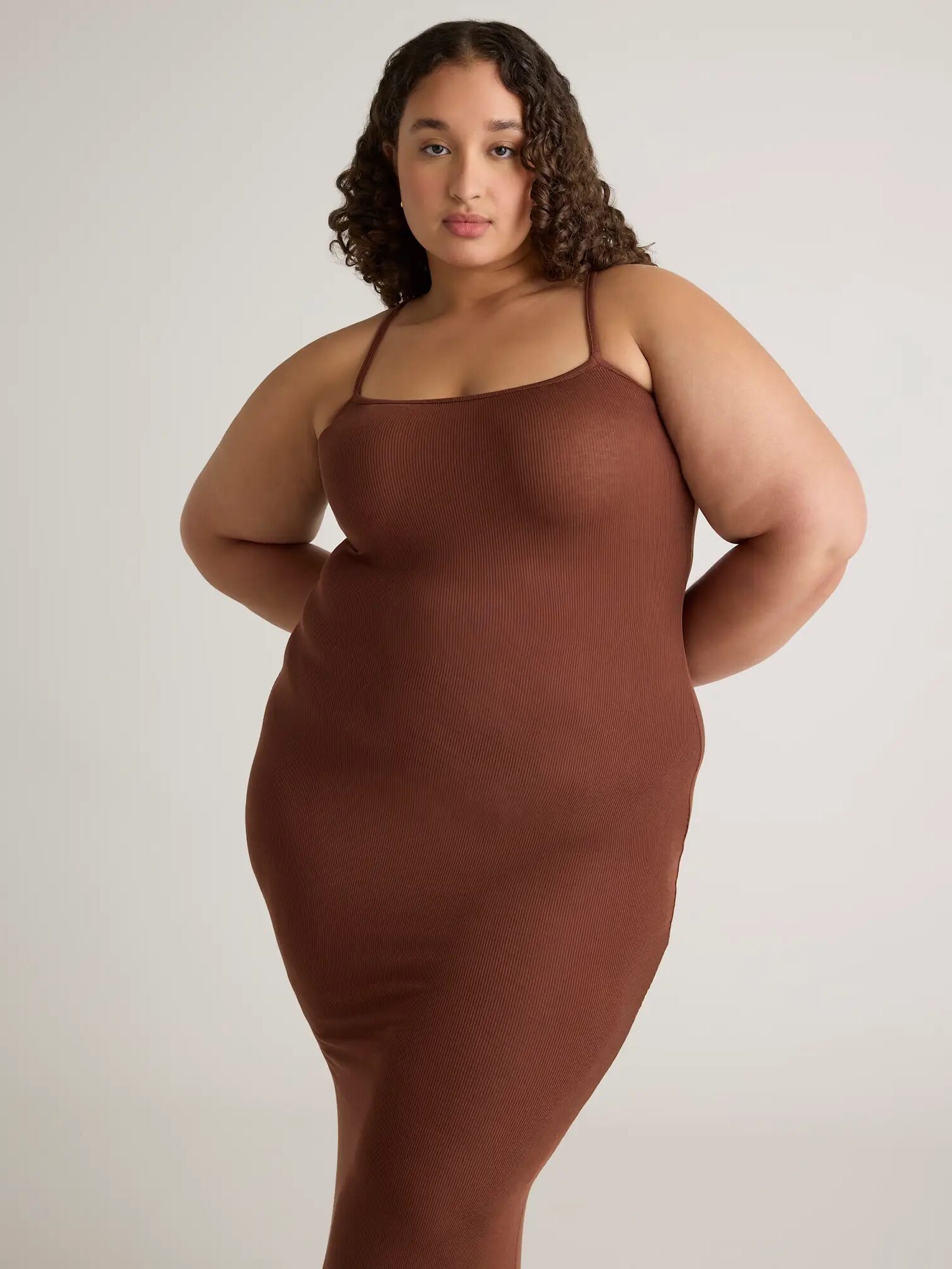 A woman with curly hair wearing a fitted, sleeveless brown dress stands against a plain light background.