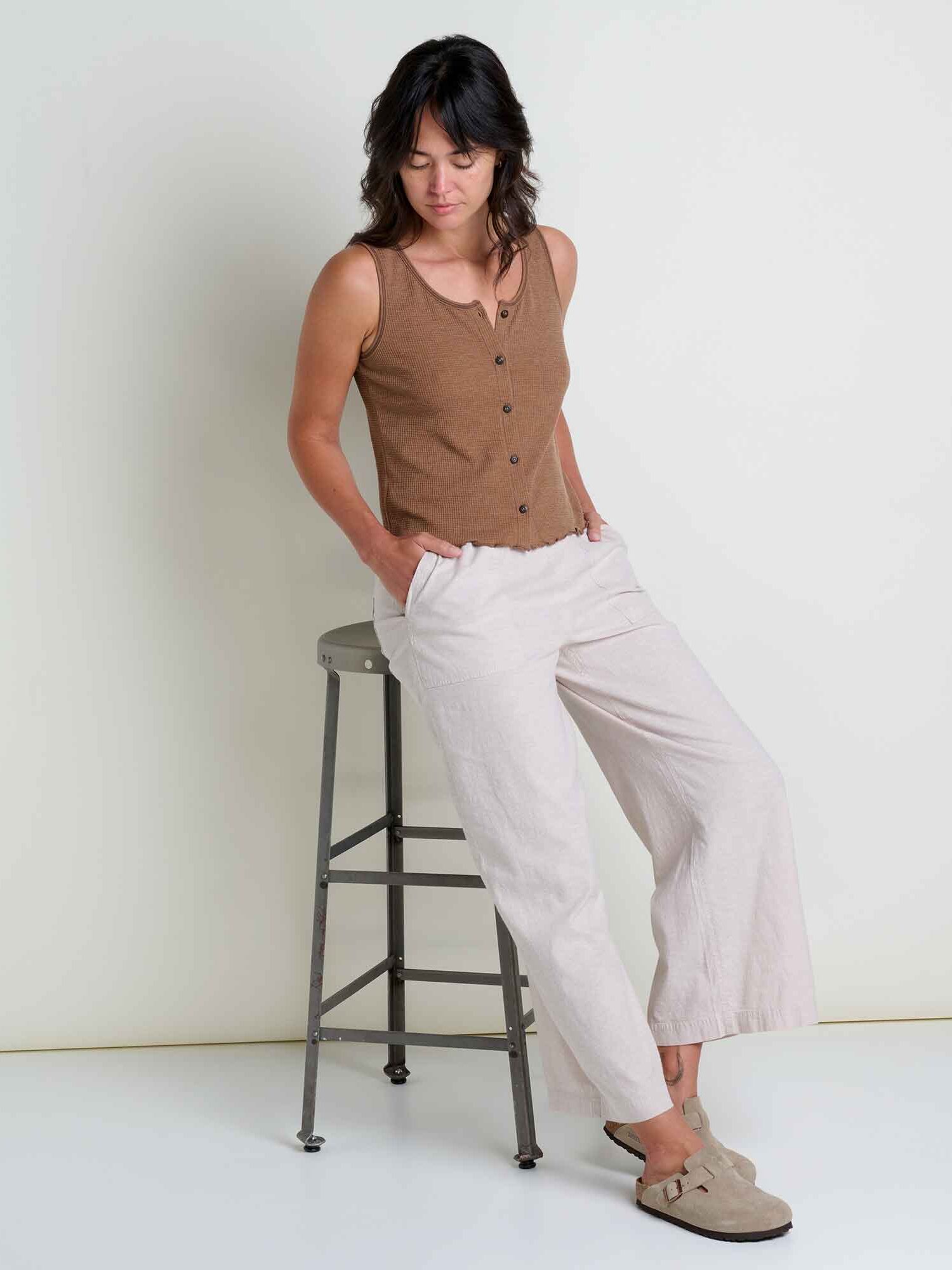 A person wearing a brown sleeveless buttoned top and light-colored wide-leg pants leans against a metal stool, looking down with hands in pockets.