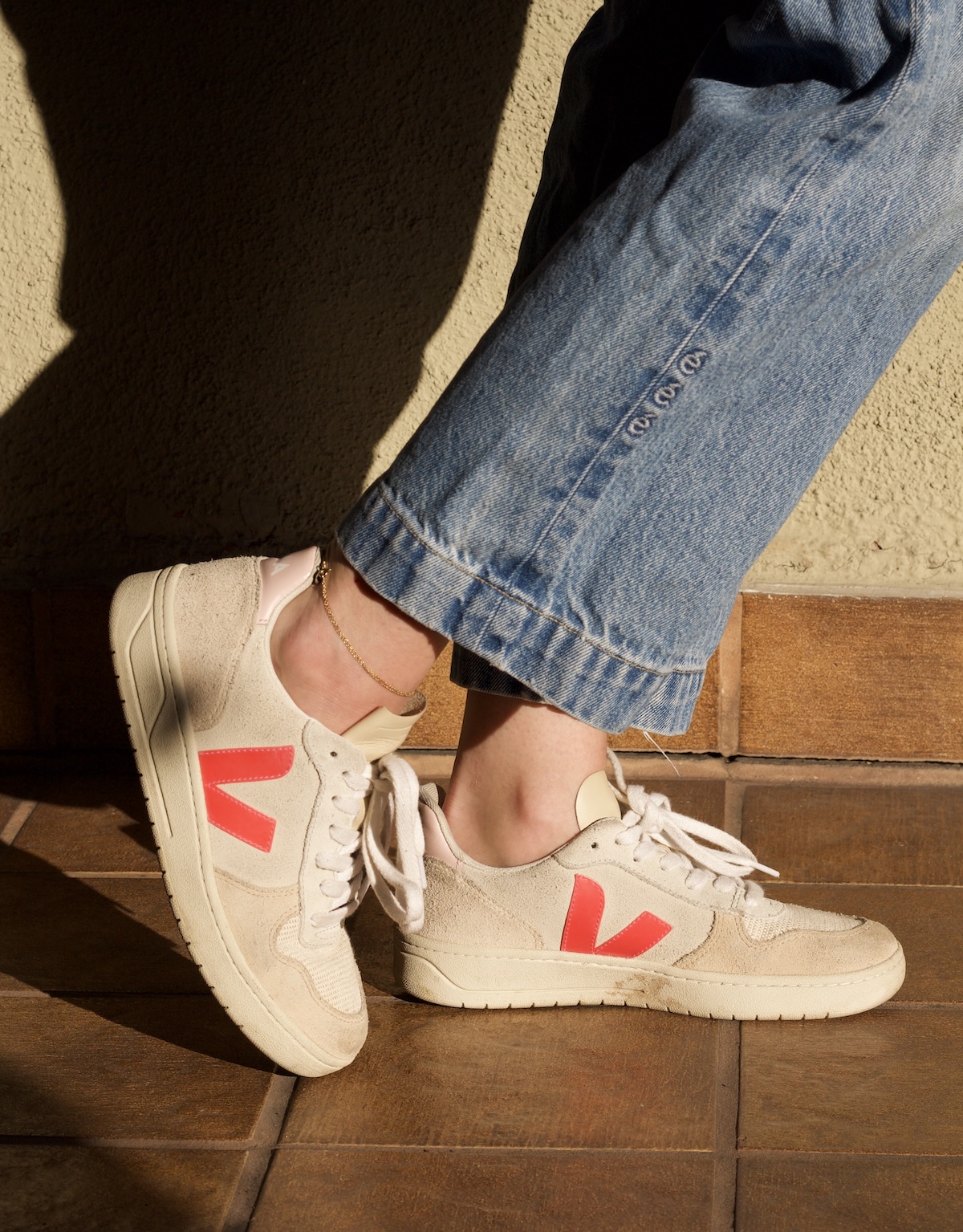 A person wearing light-wash jeans and sustainable sneakers featuring red "V" logos stands on a tiled floor in sunlight.