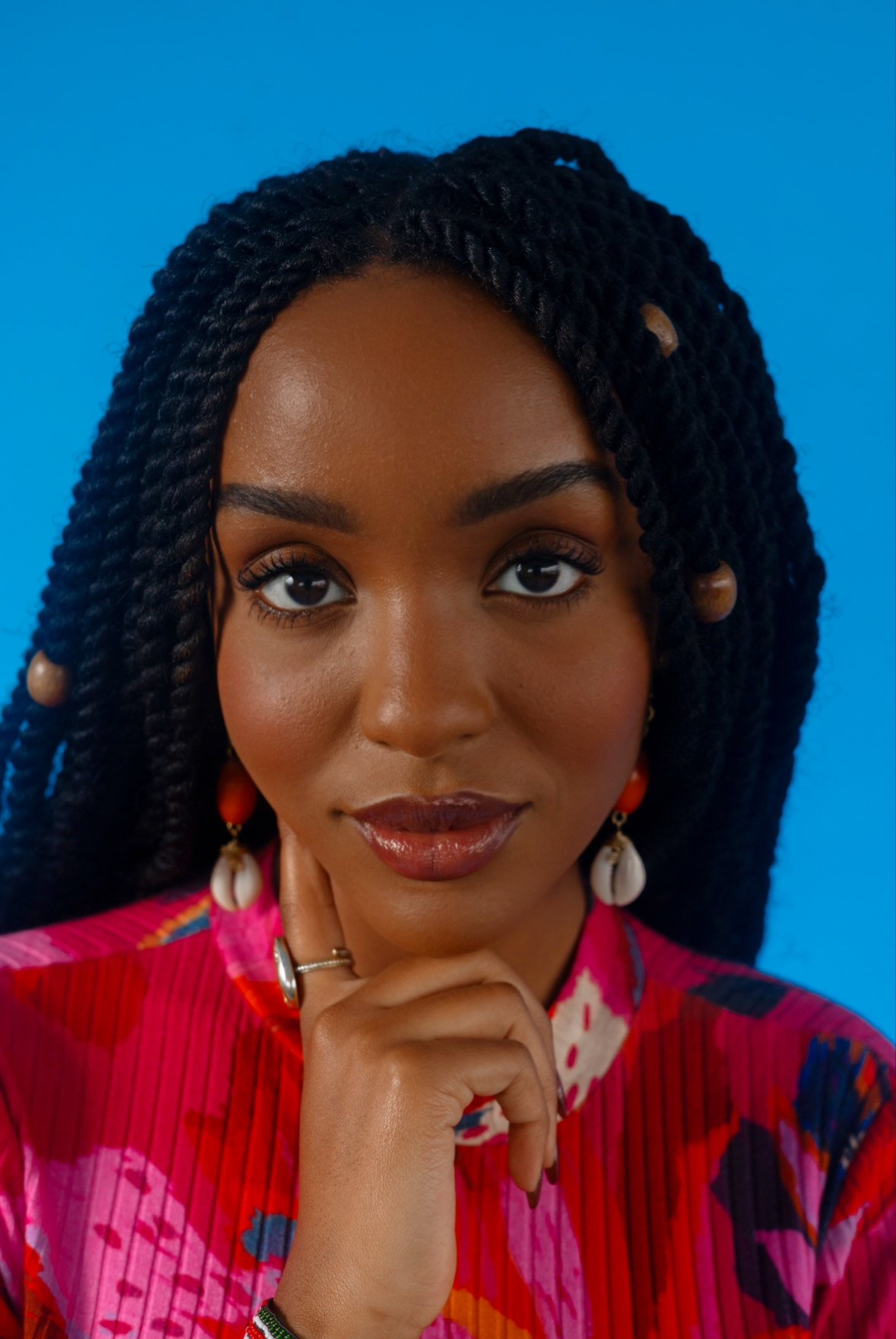 A woman with long braided hair, wearing a bright floral top, shell earrings, and a ring, poses with her hand resting under her chin against a blue background.
