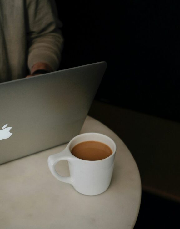 A person using a laptop sits at a white table with a cup of coffee in a white mug, organizing their tasks on Notion.