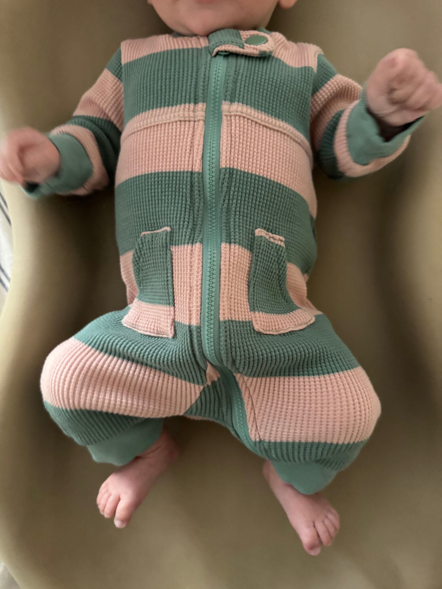 A baby wearing a pink and green striped, long-sleeve, zip-up onesie lies on a beige surface. The baby's face is not visible.
