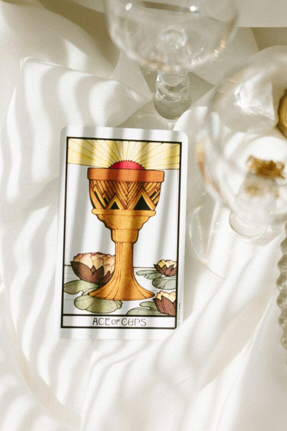 A tarot card labeled "Ace of Cups" featuring a golden chalice with water lilies, placed on a white fabric surface next to a glass.
