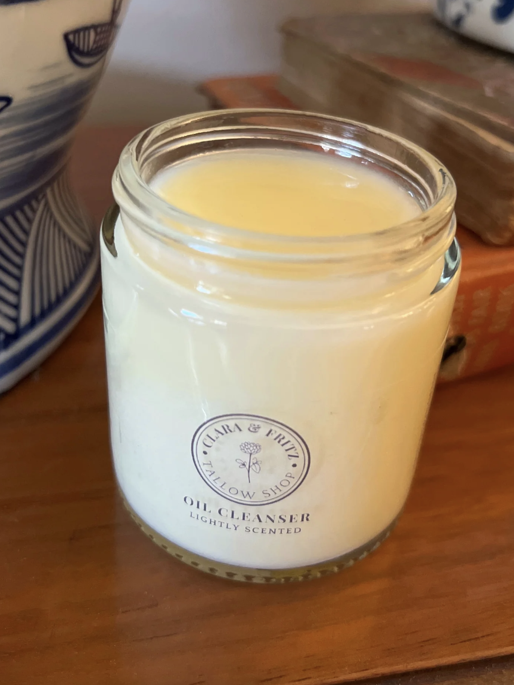 A jar of lightly scented oil cleanser is placed on a wooden surface next to decorative items and books.