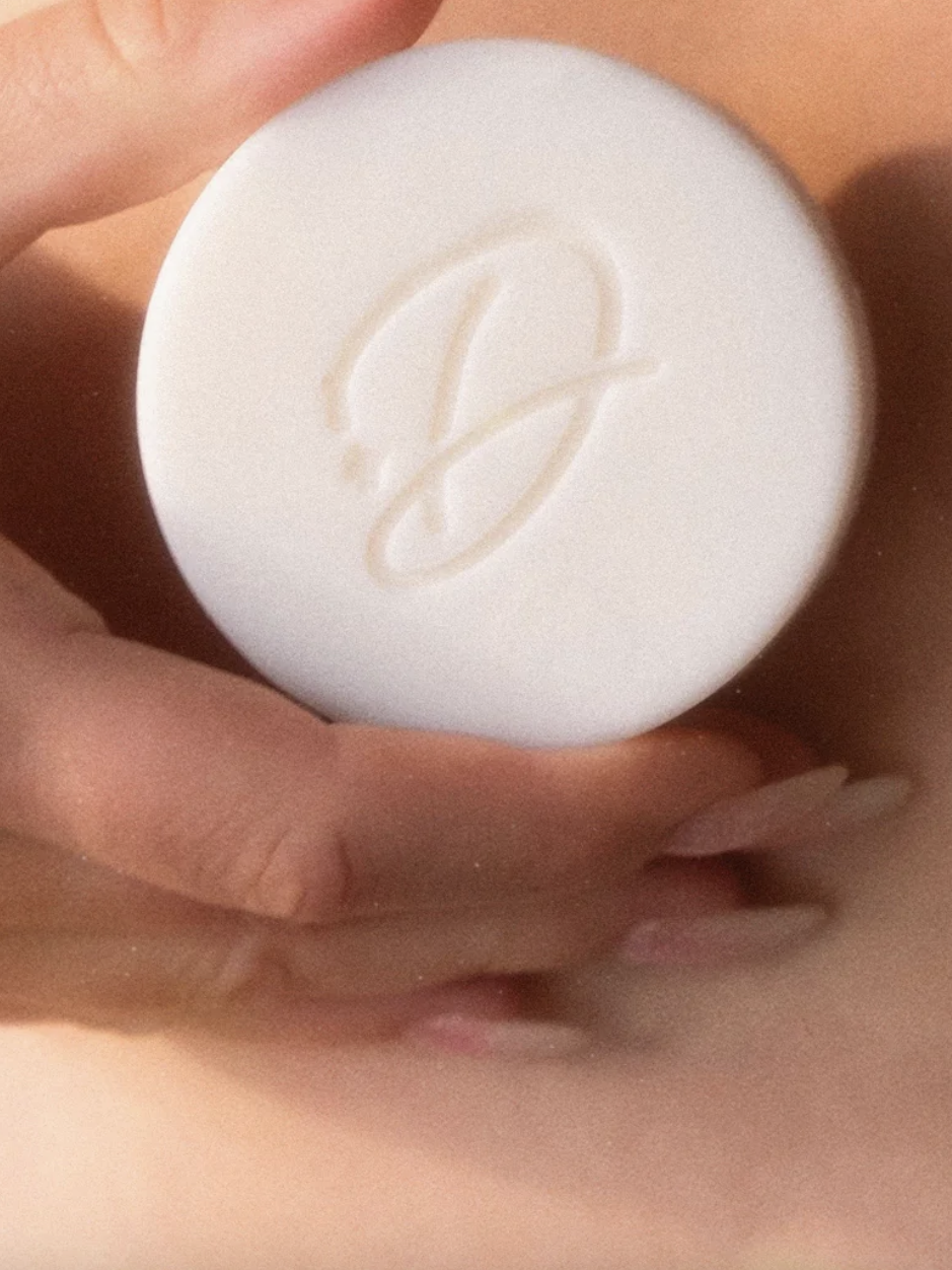 Hand holding a white round soap with an embossed letter "D" on its surface.